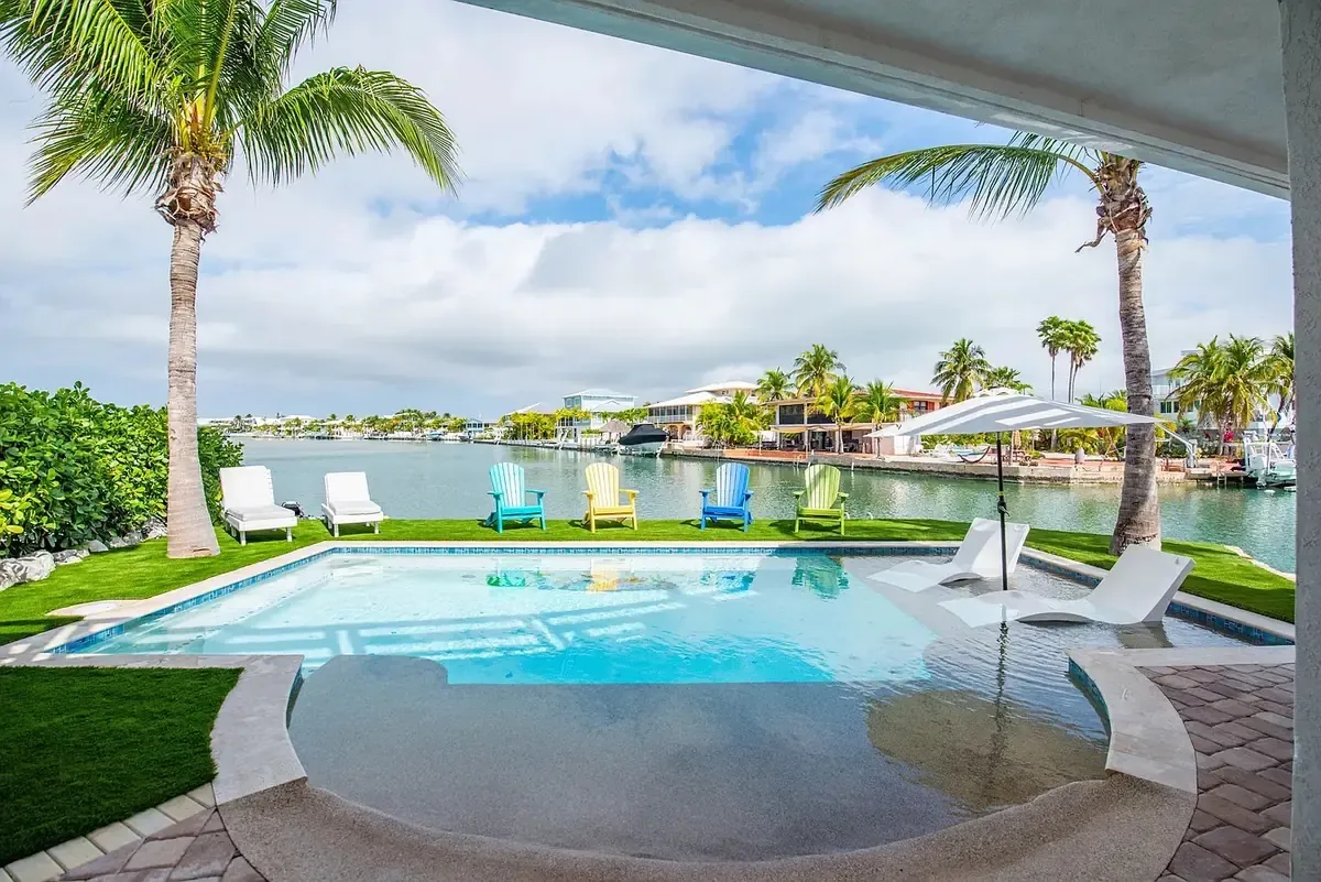 Canalfront custom pool viewed from covered patio with tanning ledge, in-water lounger, colorful Adirondack chairs, and canal views