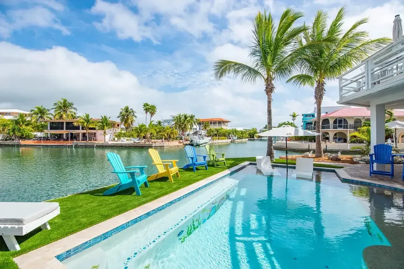 Wide angle view of canalfront custom pool with blue glass tile, in-water lounger, colorful Adirondack chairs, and waterway beyond