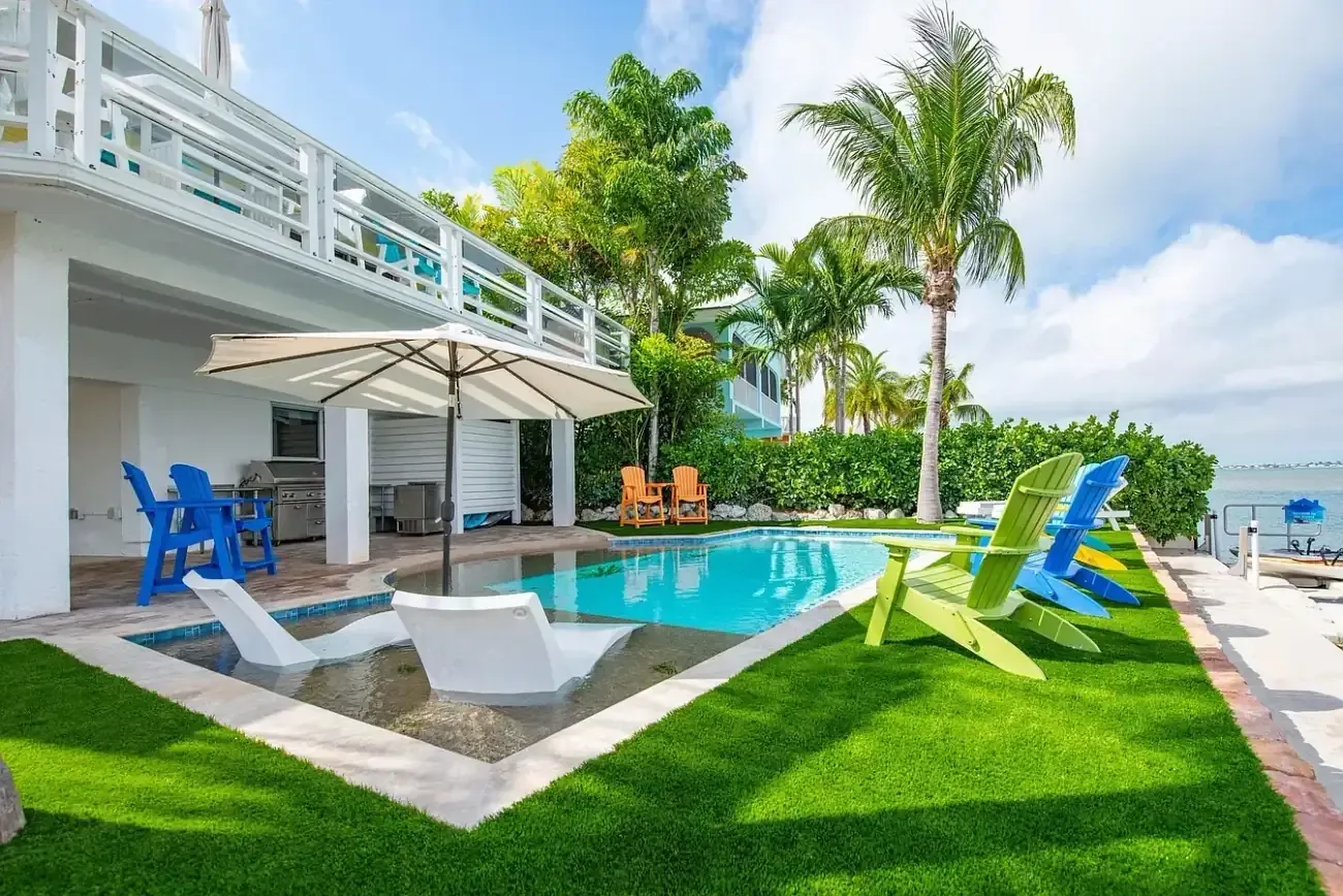 Custom pool tanning ledge with in-water loungers, patio umbrella, outdoor kitchen, and waterfront views
